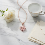 Rose quartz heart pendant necklace styled on a white marble surface with a white rose, gold-rimmed teacup, and deckle-edged paper.