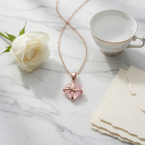 Rose quartz heart pendant necklace styled on a white marble surface with a white rose, gold-rimmed teacup, and deckle-edged paper.